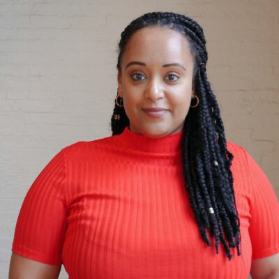 Yanet Amanuel is a Black woman in the center of the phoot. She has long braids and is wearing a bright red short-sleeved shirt.