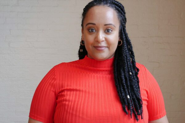 Yanet Amanuel is a Black woman in the center of the phoot. She has long braids and is wearing a bright red short-sleeved shirt.