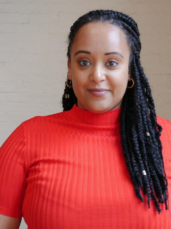 Yanet Amanuel is a Black woman in the center of the phoot. She has long braids and is wearing a bright red short-sleeved shirt.