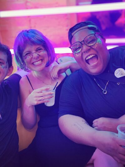 Alicia Smith on the far right is a Black, queer, non-binary person and is smiling and laughing with two friends. The background is colorful. Alicia has glitter on their face.