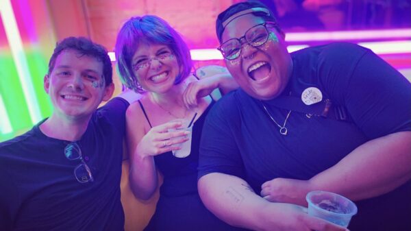 Alicia Smith on the far right is a Black, queer, non-binary person and is smiling and laughing with two friends. The background is colorful. Alicia has glitter on their face.