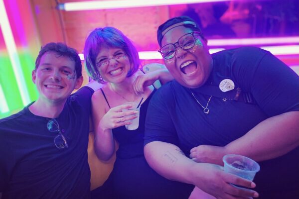 Alicia Smith on the far right is a Black, queer, non-binary person and is smiling and laughing with two friends. The background is colorful. Alicia has glitter on their face.