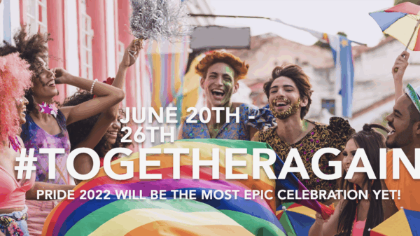 Group of people of various light to dark skin tones is laughing together holding a rainbow flag.