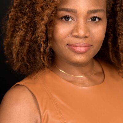 Veronica Dunlap is a Black woman with shoulder length curly hair and warm highlights. She is looking at the camera. she is wearing a sleeveless caramel-colored shirt.