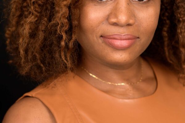 Veronica Dunlap is a Black woman with shoulder length curly hair and warm highlights. She is looking at the camera. she is wearing a sleeveless caramel-colored shirt.