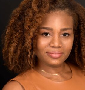 Veronica Dunlap is a Black woman with shoulder length curly hair and warm highlights. She is looking at the camera. she is wearing a sleeveless caramel-colored shirt.