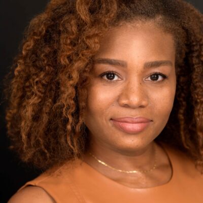 Veronica Dunlap is a Black woman with shoulder length curly hair and warm highlights. She is looking at the camera. she is wearing a sleeveless caramel-colored shirt.