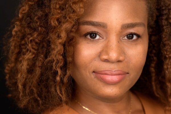 Veronica Dunlap is a Black woman with shoulder length curly hair and warm highlights. She is looking at the camera. she is wearing a sleeveless caramel-colored shirt.