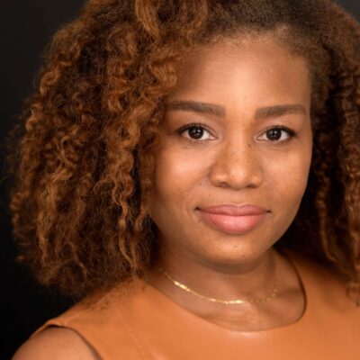 Veronica Dunlap is a Black woman with shoulder length curly hair and warm highlights. She is looking at the camera. she is wearing a sleeveless caramel-colored shirt.