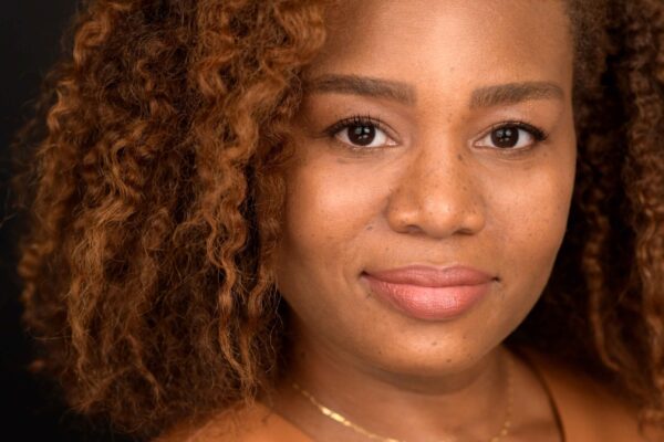 Veronica Dunlap is a Black woman with shoulder length curly hair and warm highlights. She is looking at the camera. she is wearing a sleeveless caramel-colored shirt.