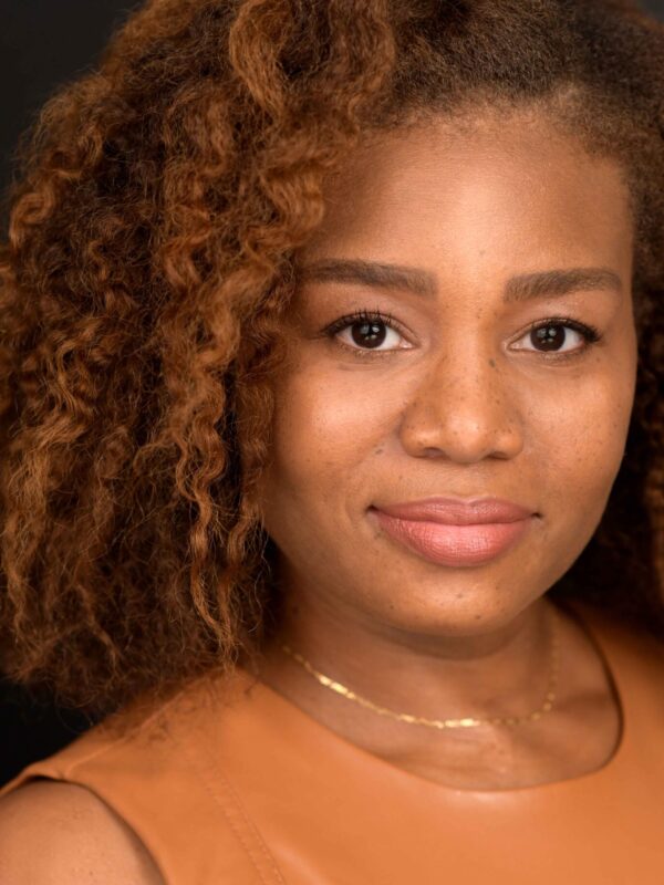 Veronica Dunlap is a Black woman with shoulder length curly hair and warm highlights. She is looking at the camera. she is wearing a sleeveless caramel-colored shirt.