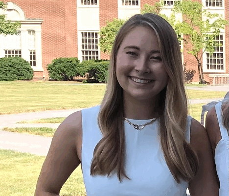Olivia Spacassi has a light skin tone, long loose wavy blondish brown hair, has on a white sleeveless top,  and is smiling at the camera. She is outside standing a little ways away in front of a brick building.