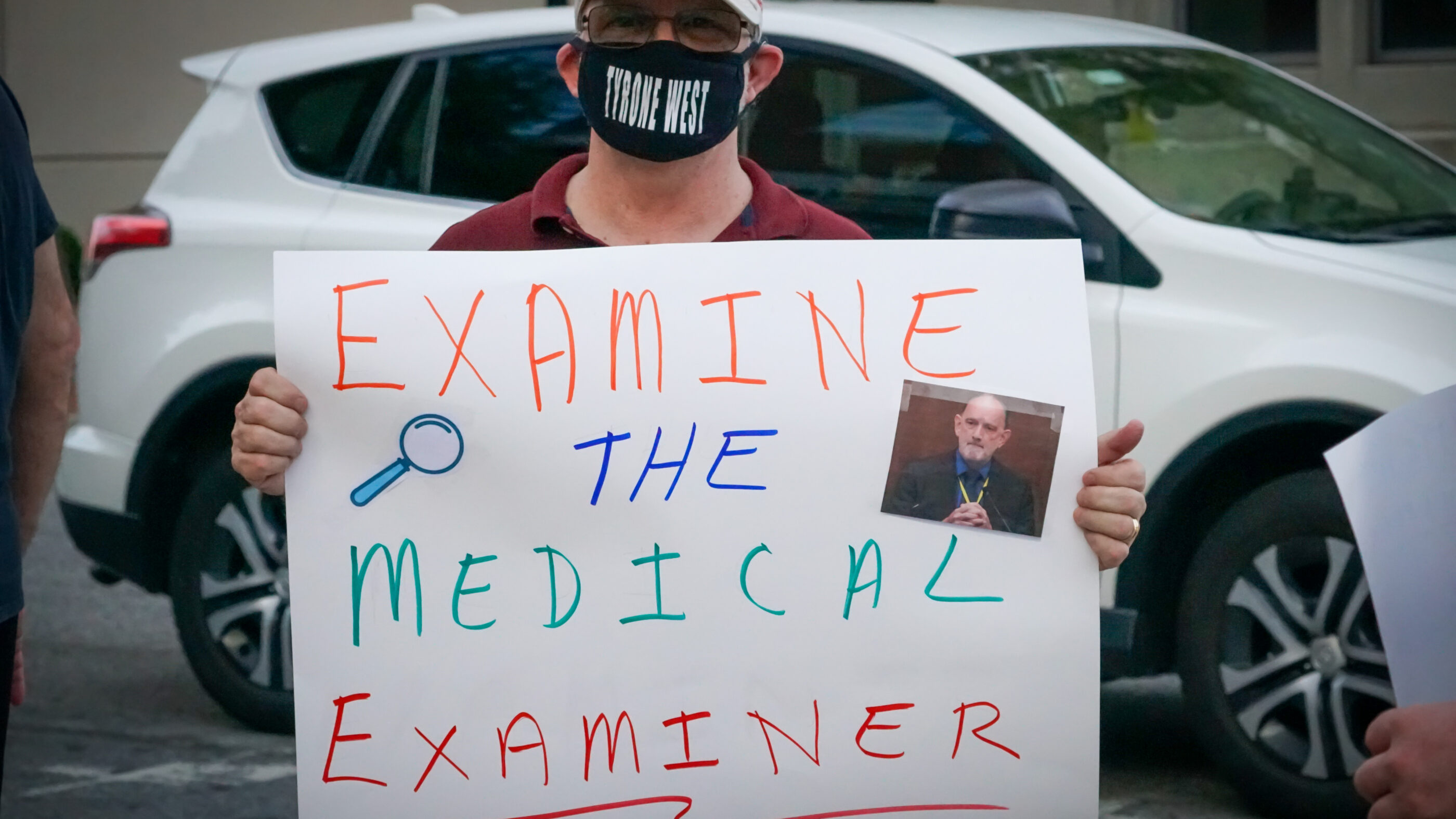 Person holds a rally sign that says, "Examine the Medical Examiner."