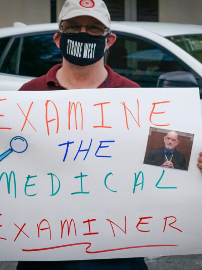 Person holds a rally sign that says, "Examine the Medical Examiner."