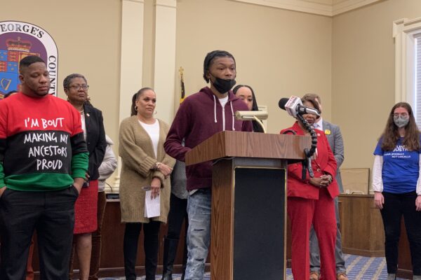 Press Conference:Group of mostly Black advocates are standing behind a podium. A young Black man is at the podium telling his story.