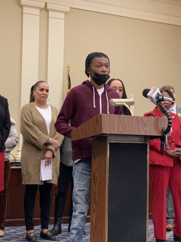 Press Conference:Group of mostly Black advocates are standing behind a podium. A young Black man is at the podium telling his story.