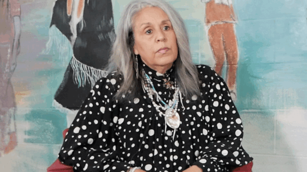 Cedarville Band of the Piscataway Indians Tribal Chairwoman Standing on the Rock Natalie Proctor is sitting in front of a mural at their cultural center. She has medium length gray hair and is wearing Indigenous necklaces.