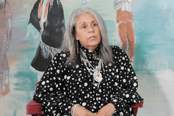 Cedarville Band of the Piscataway Indians Tribal Chairwoman Standing on the Rock Natalie Proctor is sitting in front of a mural at their cultural center. She has medium length gray hair and is wearing Indigenous necklaces.