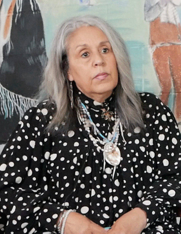 Cedarville Band of the Piscataway Indians Tribal Chairwoman Standing on the Rock Natalie Proctor is sitting in front of a mural at their cultural center. She has medium length gray hair and is wearing Indigenous necklaces.