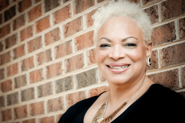 Dana Vickers Shelley is a Black woman with warm light brown skin, blond short hair, and is smiling at the camera. She is standing in front of a brick wall.