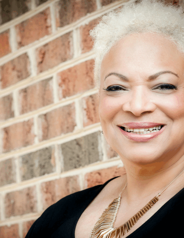 Dana Vickers Shelley is a Black woman with warm light brown skin, blond short hair, and is smiling at the camera. She is standing in front of a brick wall.