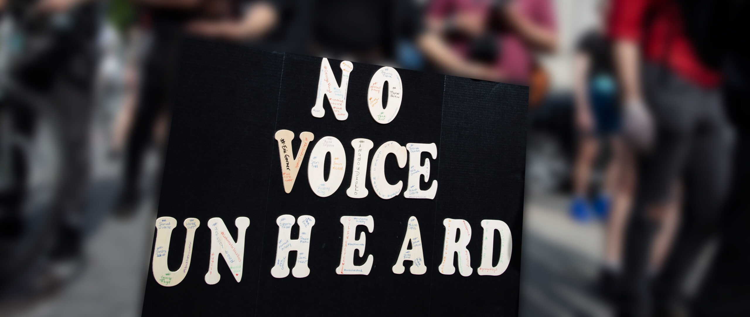 A person's hands are holding a large protest sign that says, "No Voice Unheard." The background is blurred out but you can see that there are people at a protest. Courtesy credit: Molly Kaplan, ACLU.