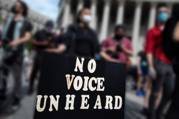 A person's hands are holding a large protest sign that says, "No Voice Unheard." The background is blurred out but you can see that there are people at a protest. Courtesy credit: Molly Kaplan, ACLU.