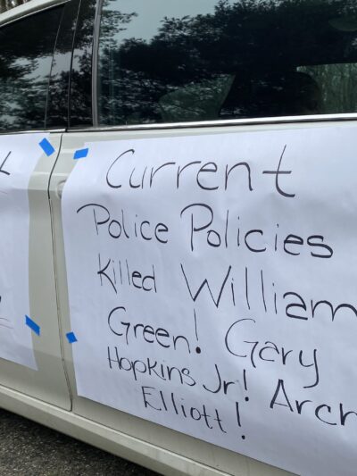 Rally signs are taped to the side of a car. They say, "Public Input = Public Safety. Current Police Practiceds Killed Willian Green, Gray Hopkins, Jr., Archie Elliot!
