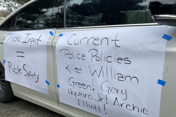 Rally signs are taped to the side of a car. They say, "Public Input = Public Safety. Current Police Practiceds Killed Willian Green, Gray Hopkins, Jr., Archie Elliot!