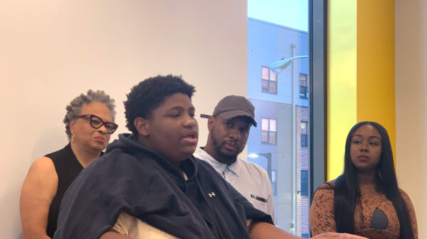 A Black student is speaking at the podium at a press conference for the launch of the National Youth Justice Action Month in Baltimore City. Behind the student are Dana Vickers Shelley, Dayvon Love, and another person from the No Boundaries Coalition.