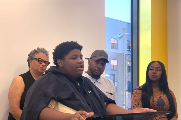 A Black student is speaking at the podium at a press conference for the launch of the National Youth Justice Action Month in Baltimore City. Behind the student are Dana Vickers Shelley, Dayvon Love, and another person from the No Boundaries Coalition.