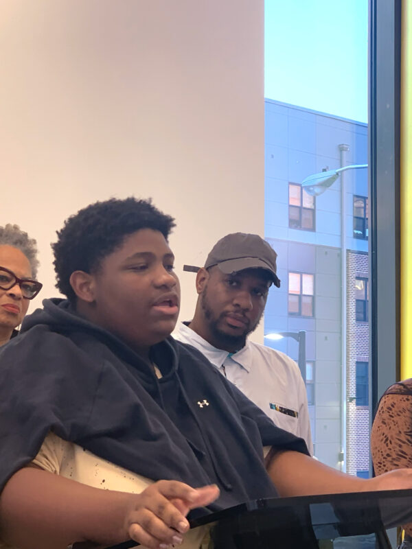 A Black student is speaking at the podium at a press conference for the launch of the National Youth Justice Action Month in Baltimore City. Behind the student are Dana Vickers Shelley, Dayvon Love, and another person from the No Boundaries Coalition.
