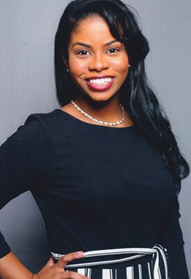 Rebecca Phillips is a Black woman who is looking and smiling at the camera. She has long wavy black hair, has caramel brown skin, is wearing red lipstick and a long-sleeve dress with black top and black and white striped skirt. She has a hand on her hip.