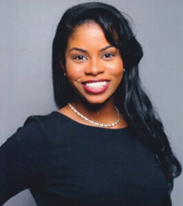 Rebecca Phillips is a Black woman who is looking and smiling at the camera. She has long wavy black hair, has caramel brown skin, is wearing red lipstick and a long-sleeve dress with black top and black and white striped skirt. She has a hand on her hip.