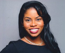 Rebecca Phillips is a Black woman who is looking and smiling at the camera. She has long wavy black hair, has caramel brown skin, is wearing red lipstick and a long-sleeve dress with black top and black and white striped skirt. She has a hand on her hip.