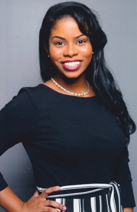 Rebecca Phillips is a Black woman who is looking and smiling at the camera. She has long wavy black hair, has caramel brown skin, is wearing red lipstick and a long-sleeve dress with black top and black and white striped skirt. She has a hand on her hip.