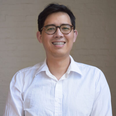 Nick Taichi Steiner is a Japanese American man with short dark hair and is wearing glasses. He is looking at the camera and smiling. he has a white button-up shirt on and a light brick wall behind him.