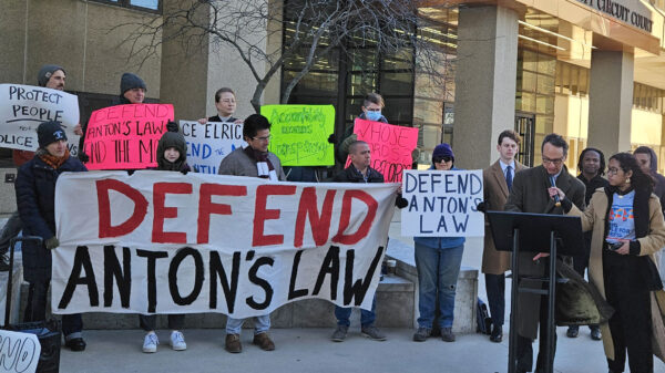 Group of advocates gather in front of the Montgomery County Circuit Courthouse at a press conference to defend Anton's Law. There is a person at a podium and about 20 advocates holding banners and signs.
