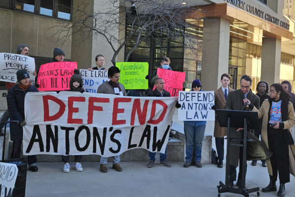 Group of advocates gather in front of the Montgomery County Circuit Courthouse at a press conference to defend Anton's Law. There is a person at a podium and about 20 advocates holding banners and signs.