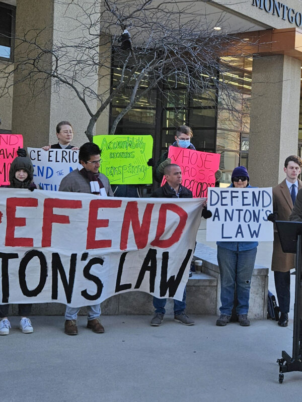 Group of advocates gather in front of the Montgomery County Circuit Courthouse at a press conference to defend Anton's Law. There is a person at a podium and about 20 advocates holding banners and signs.