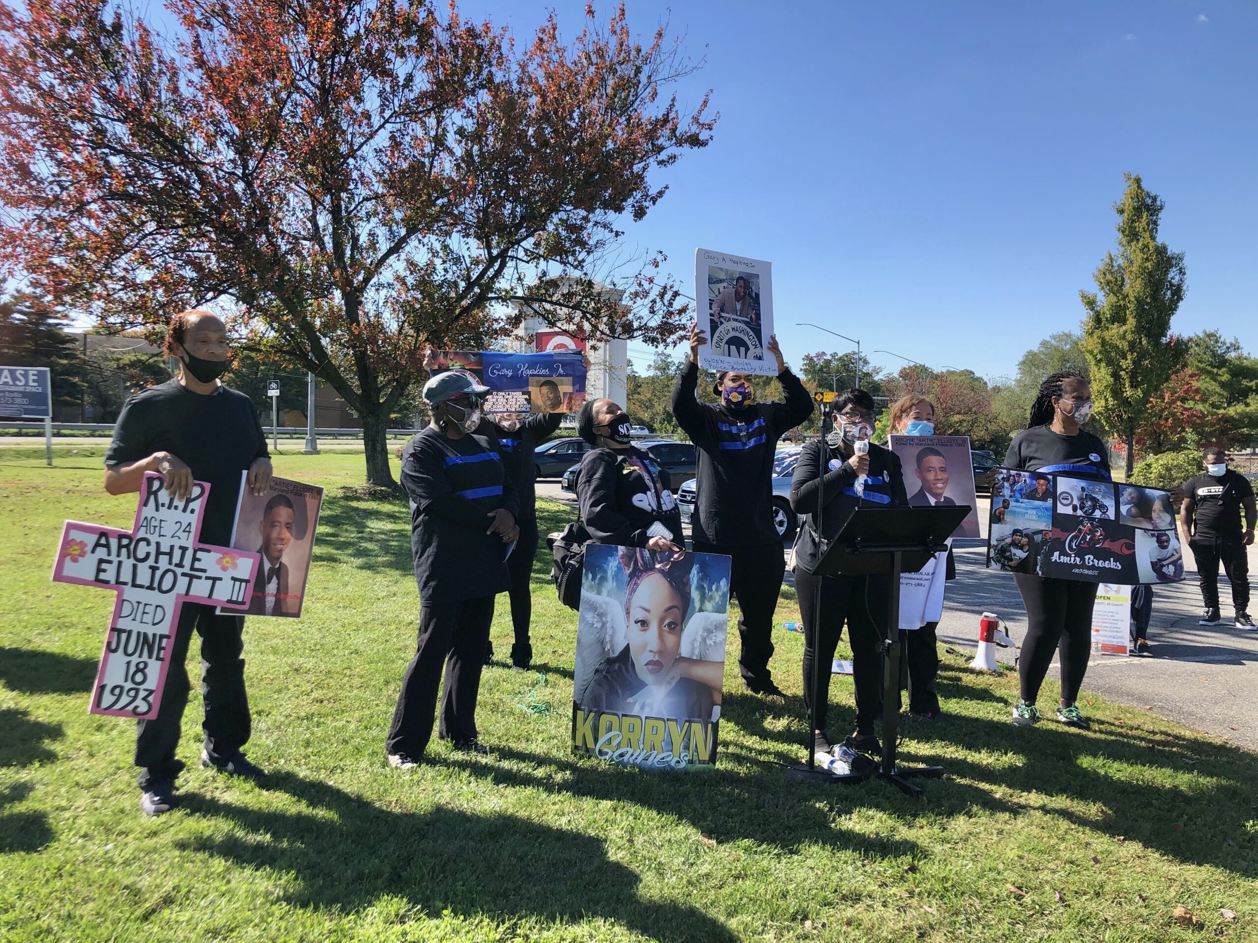 Prince George's County rally photo for mothers of Black peoole killed by police. A group of people are standing in a park holding signs with people's names who were killed.