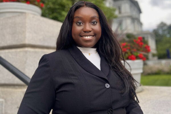 Chinyere Okonkwo is a Black woman with long dark hair. She is standing outside in front of a building with flowers and green plants. She is wearing a black blazer and is smiling at the camera.
