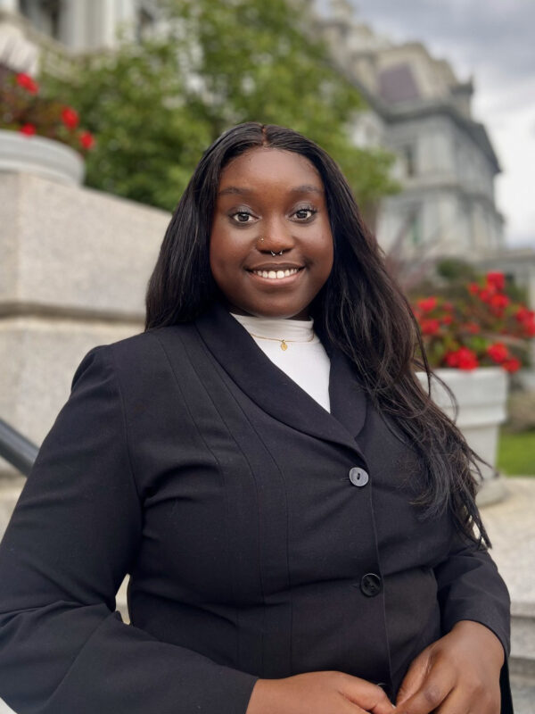 Chinyere Okonkwo is a Black woman with long dark hair. She is standing outside in front of a building with flowers and green plants. She is wearing a black blazer and is smiling at the camera.