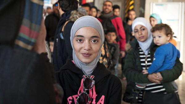 A Muslim person is looking at the camera and has a slight smile on their face. They are wearing a hijab. There is a Muslim parent holding a child in the background.