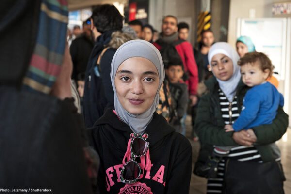 A Muslim person is looking at the camera and has a slight smile on their face. They are wearing a hijab. There is a Muslim parent holding a child in the background.
