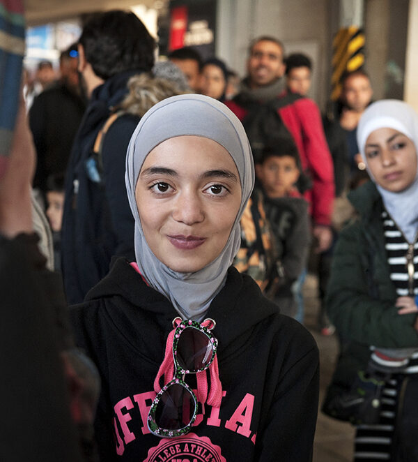 A Muslim person is looking at the camera and has a slight smile on their face. They are wearing a hijab. There is a Muslim parent holding a child in the background.