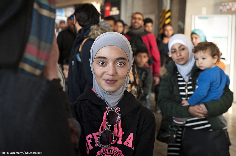 A Muslim person is looking at the camera and has a slight smile on their face. They are wearing a hijab. There is a Muslim parent holding a child in the background.