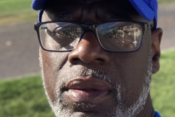 Ransom Watkins is a Black man wearing a blue baseball hat, glasses, has a short gray beard, and is wearing a blue shirt. He is standing outside.