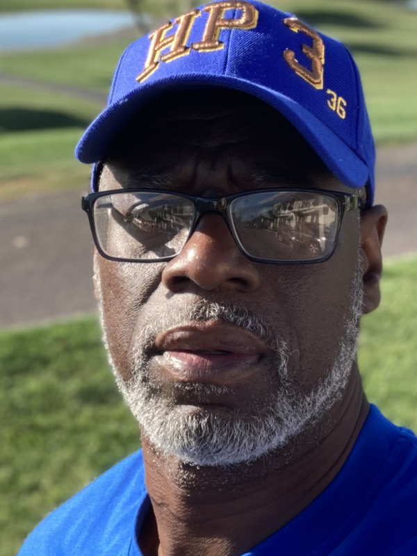 Ransom Watkins is a Black man wearing a blue baseball hat, glasses, has a short gray beard, and is wearing a blue shirt. He is standing outside.