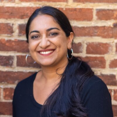 Sonia Kumar is a Middle Eastern woman with warm mediu brown skin tone. She is smiling at the camera and has long black hair pulled back in a low pony tail. She is wearing hoop earrings. She is standing in front of brick wall.